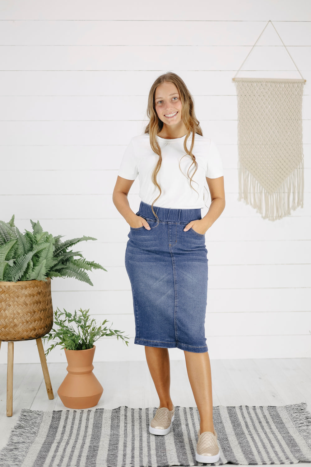 A woman standing in a room with a white wall and decorative hanging behind her, wearing a white t-shirt and a mid-calf length indigo denim skirt with pockets.