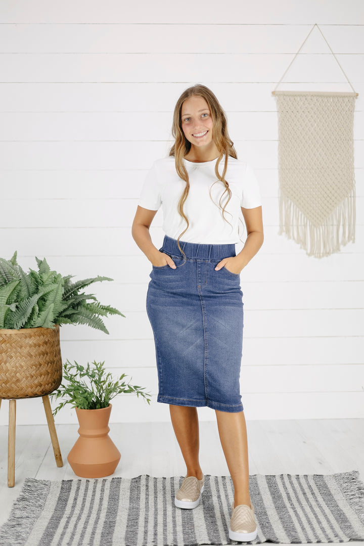A woman standing in a room with a white wall and decorative hanging behind her, wearing a white t-shirt and a mid-calf length indigo denim skirt with pockets.