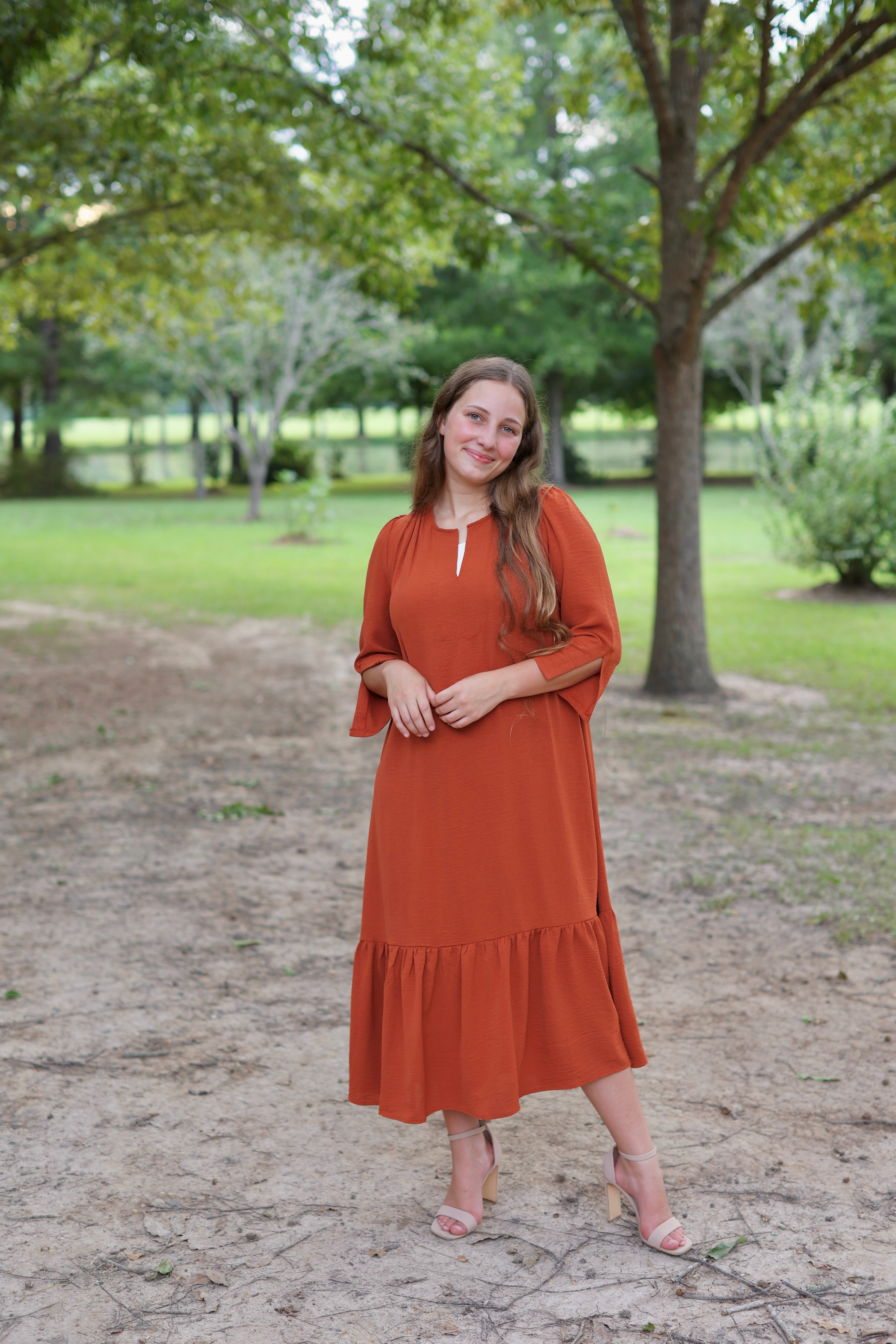 Woman in a rust-colored dress standing in a park with trees and grass in the background