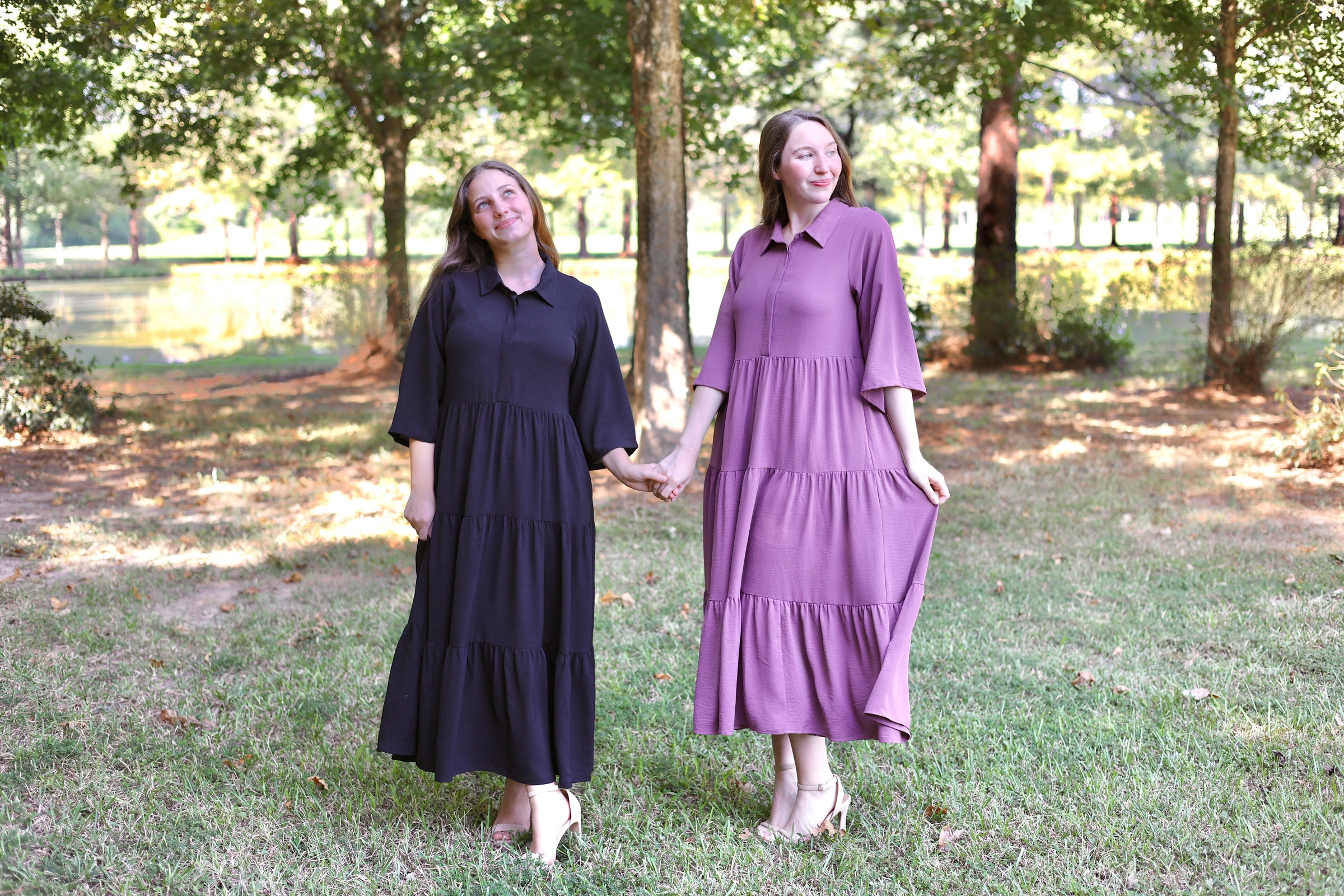 Two women standing outdoors holding hands, one in a black dress and the other in a purple dress.